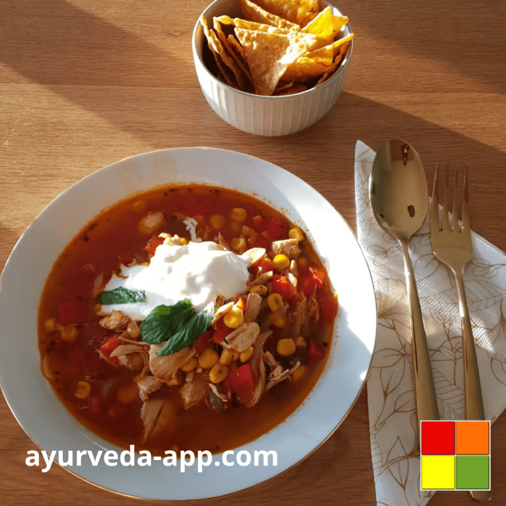 Photo of Ayurveda Chicken Tortilla Soup served in a white bowl with golden rims. there's also another bowl of tortilla chips on the side. Next to the soup is a napkin decorated with a fork and spoon.