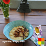 Photo of date and oat porridge served in a blue bowl with brown rims. Next to the bowl is a spoon on top of a flowered napkin. Behind is a vase of flowers.