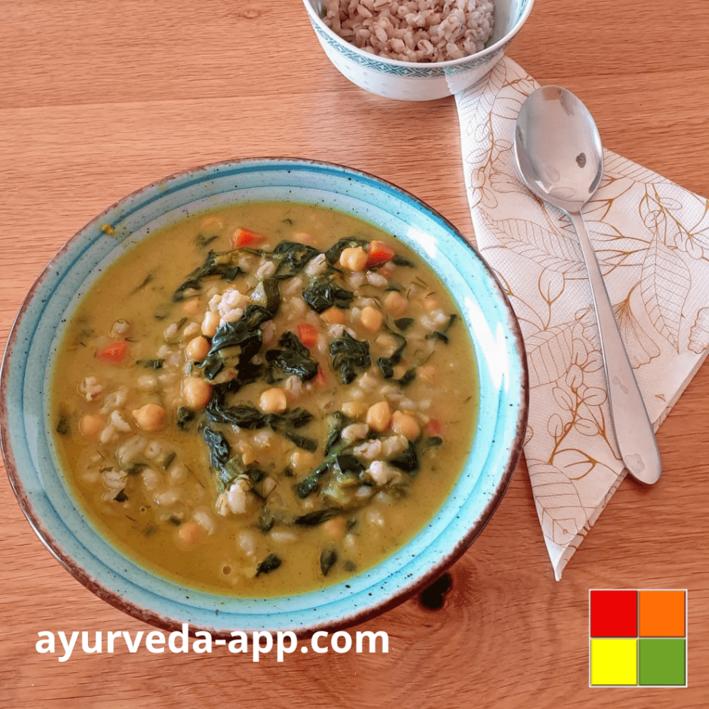 Photo of a blue bowl of Chickpea spinach soup with barley. Next to the bowl is a decorated napkin with a spoon on top, behind it is a small bowl of barley.