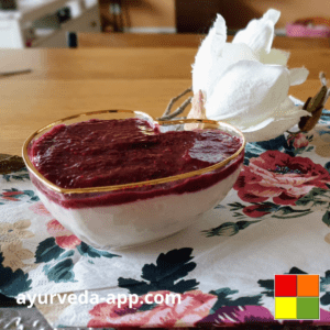 Photo of a golden-rimmed heart-shaped bowl of Coconut pudding with raspberry and chia. The bowl is on top of a floral decorated napkin. Behind, there is a white flower.