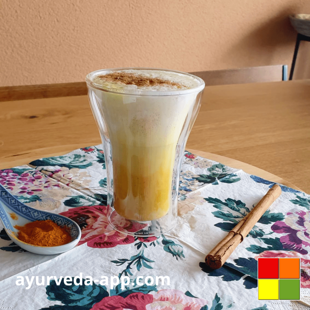 Photo of Golden Milk/Turmeric Latte served in a double glass cup on top of a decorated napkin. On the side, there is cinnamon and turmeric to decorate.