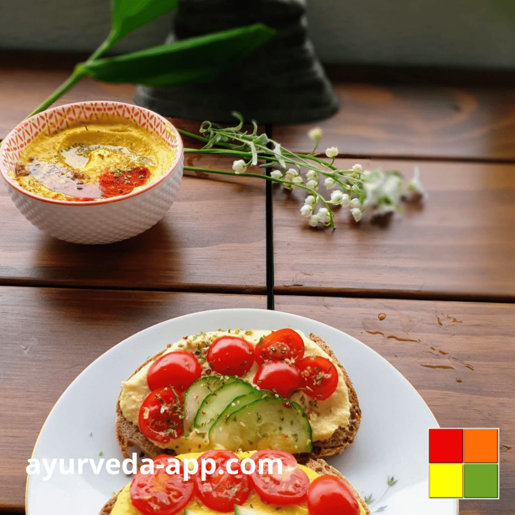 Photo of a plate with two slices of bread with Ayurvedic Curry Hummus, tomatoes and cucumbers. There is also a bowl with the hummus. The environment is decorated with leaves and flowers.