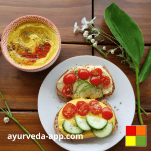 Photo of a plate with two slices of bread with Ayurvedic Curry Hummus, tomatoes, and cucumbers. There is also a bowl with the hummus. The environment is decorated with leaves and flowers.