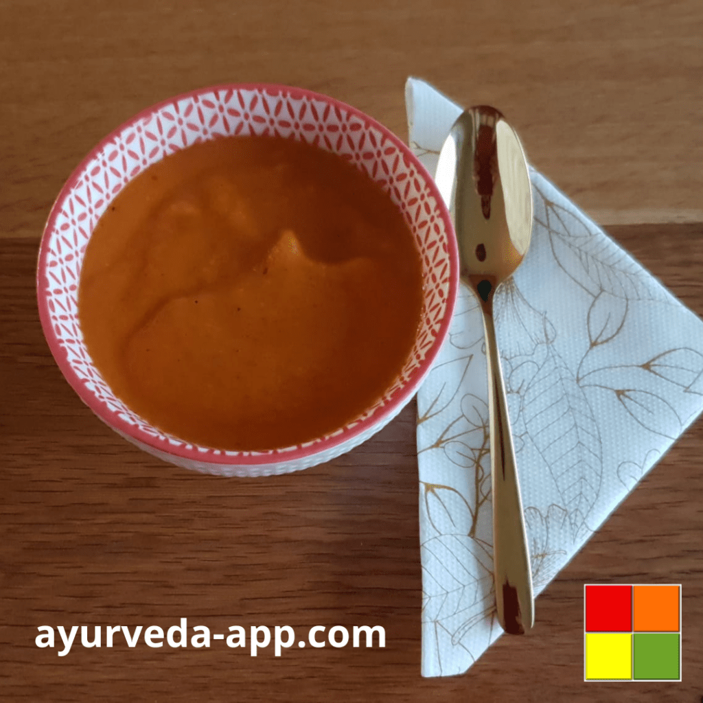 Photo of Ginger carrot soup served in a white bowl with red accents. Next to the bowl is a spoon on top of a decorated napkin.