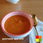 Photo of Ginger carrot soup served in a white bowl with red accents. Next to the bowl is a spoon on top of a decorated napkin.