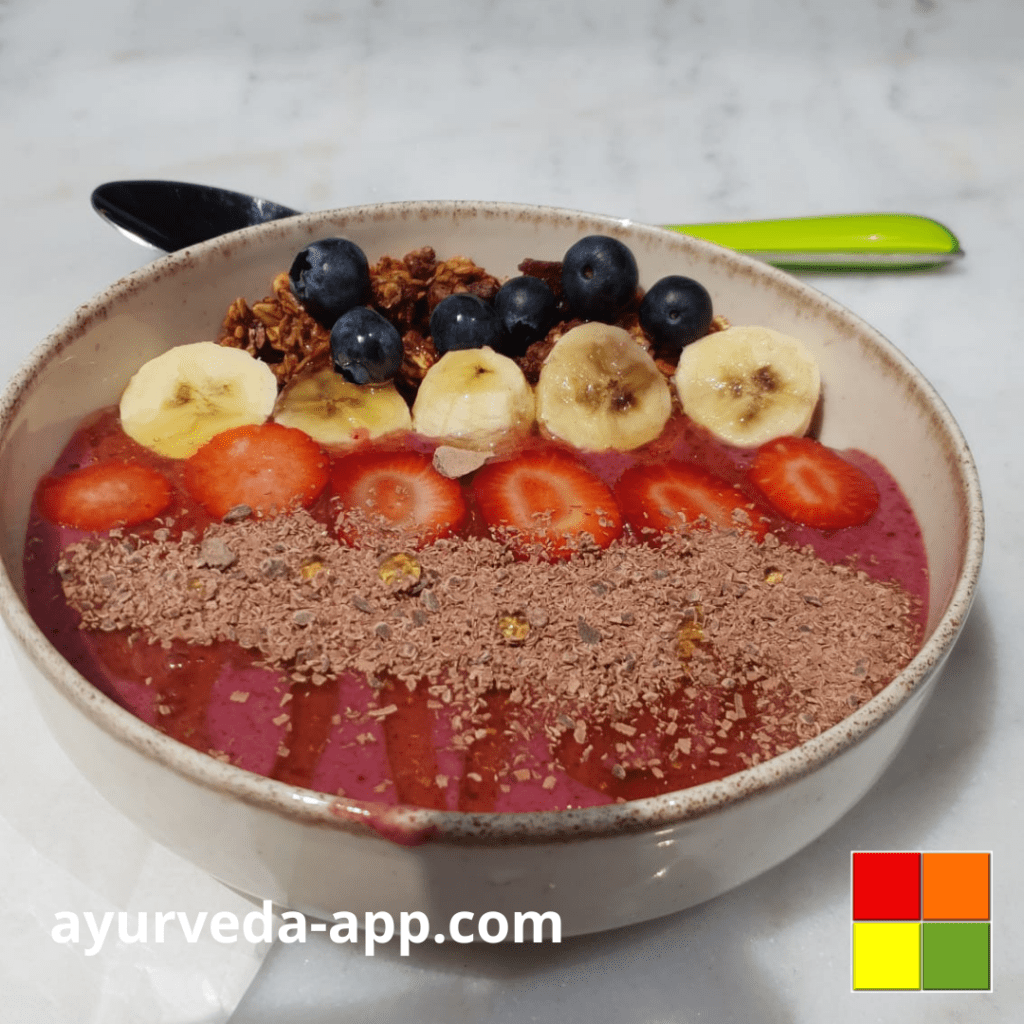 Photo of an Ayurvedic smoothie bowl. The recipe is decorated with strawberries, bananas, granola, blueberries, and cacao nibs. Behind the bowl is a green spoon.