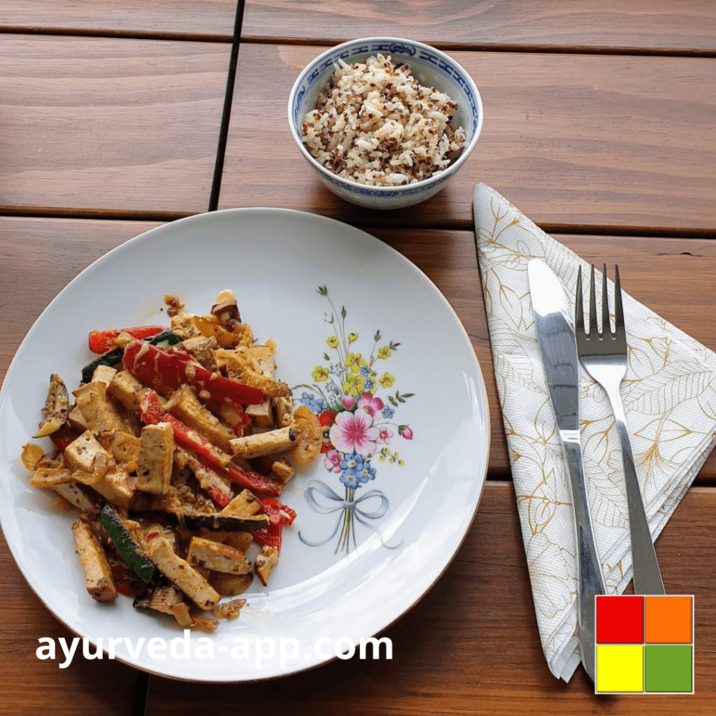 Photo of Tofu with vegetables and coconut sauce. The dish is served on a white plate decorated with flowers. Behind the plate, there is a bowl with a mix of rice and quinoa and beside it, there is a fork and knife on top of a decorated napkin.