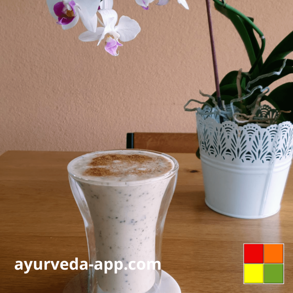 Photo of Cashew Banana Smoothie served in a double glass tumbler. In the background there is a vase of orchids.