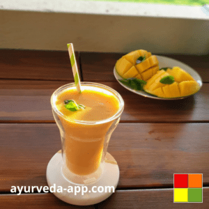 Photo of a double glass cup with mango lassi and a white and green paper cane, along with a plate of mangoes.