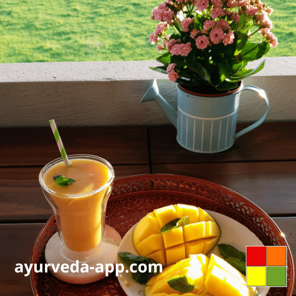Photo of a double glass cup with mango lassi and a white and green paper straw. The glass is on a decorated red tray along with a plate of mangoes. Behind the tray is a blue vase in the shape of a watering can with pink flowers.
