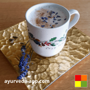 Photo of a Moon Milk mug. The mug is white with details of leaves and flowers. The mug is on top of a golden plate. Next to the mug are lavender flowers to decorate.