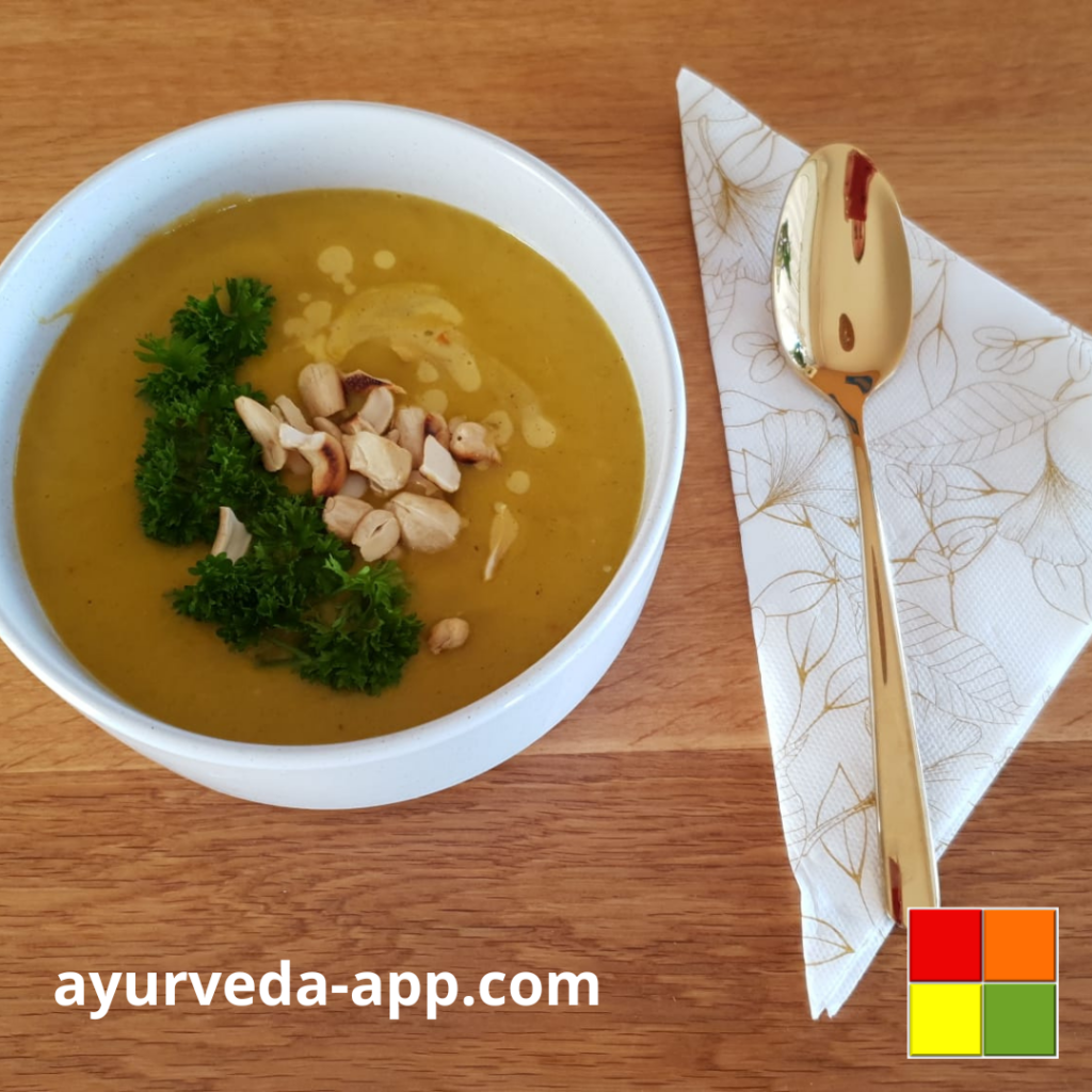 Photo of Cashew pumpkin leek soup served in a white bowl, topped with cashews and fresh herbs for garnish. Next to the bowl is a golden spoon on top of a decorated napkin.