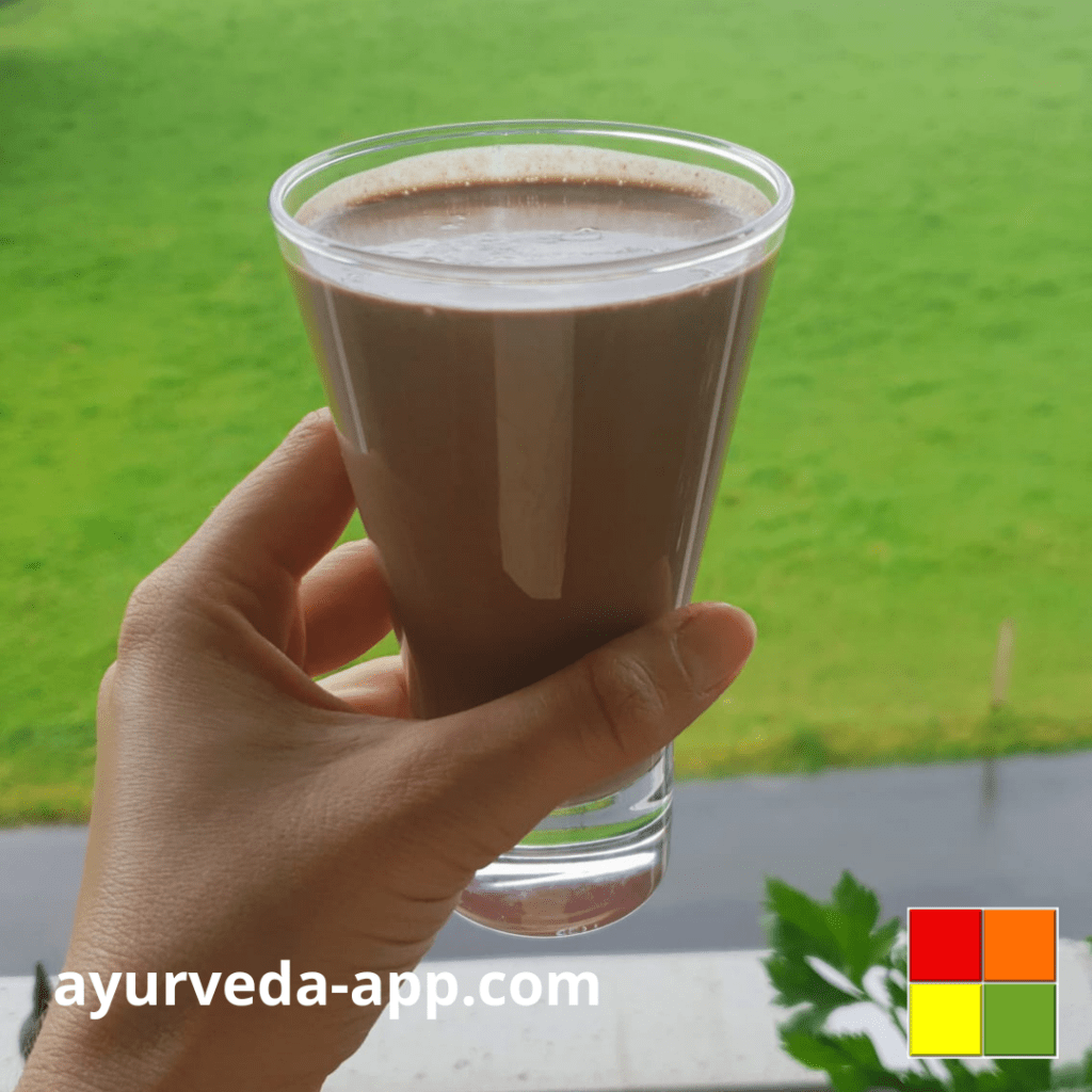 Photo of a hand holding a glass of Pecan chocolate smoothie. In the background of the image there is grass.