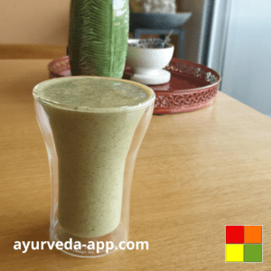 Photo of a double glass glass of glowy skin smoothie. In the background there is a green jug and a white bowl on top of a decorated red tray.