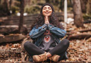 Photo of a smiling woman sitting with legs crossed on the floor, in the position for Yoga, meditation and pranayama. Hands with palms together, just below the chin. She is in nature, sitting around dry leaves and has a cat next to her.