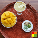 In the image there is a glass of the result of the tridosha balancing smoothie recipe, in a glass cup with a glass straw. The glass is on top of a decorated red tray and next to it there is also a bowl with diced mango and a small plate with the three powdered supplements used in the recipe.