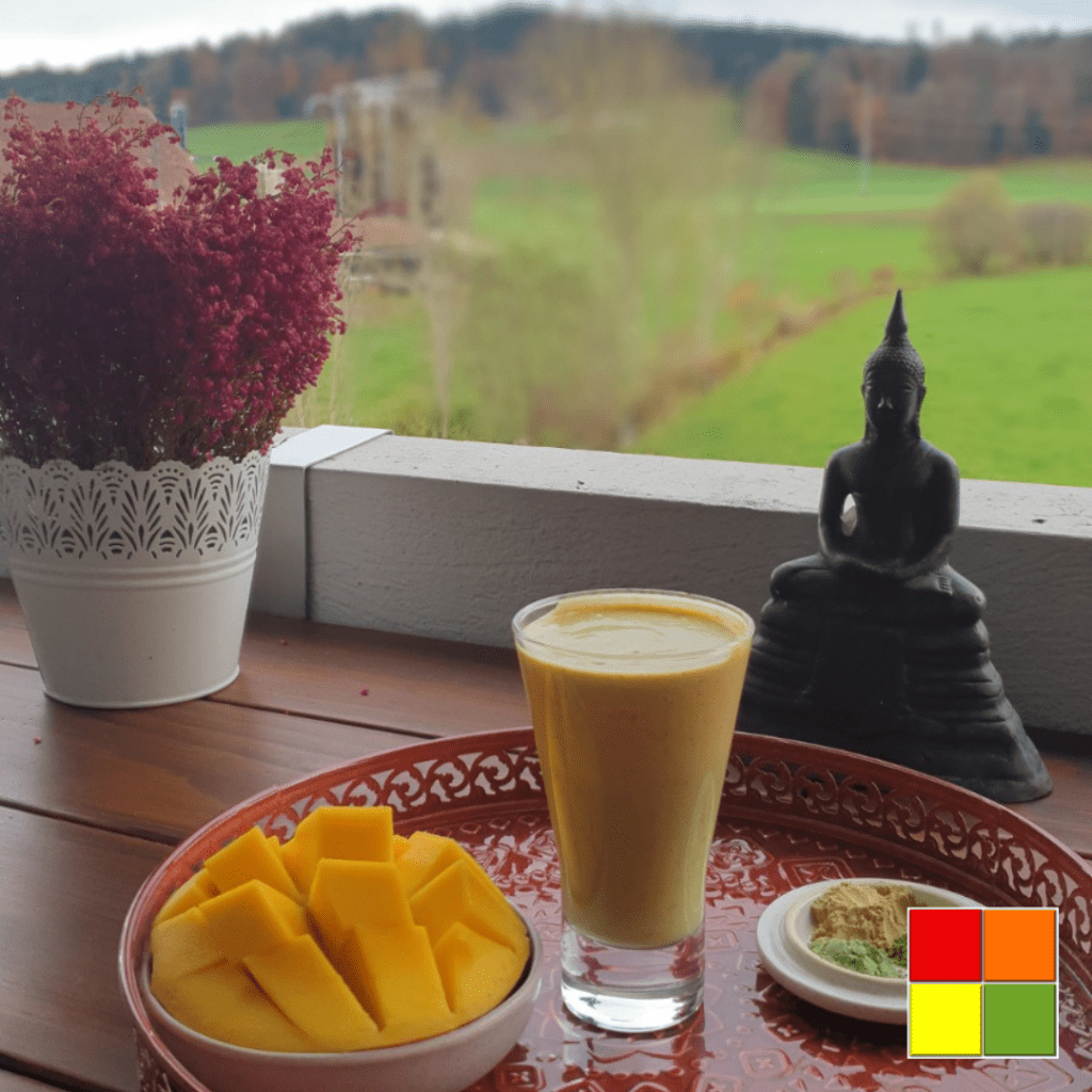 In the image there is a glass of the result of the tridosha balancing smoothie recipe, in a glass cup with a glass straw. The glass is on top of a decorated red tray and next to it there is also a bowl with diced mango and a small plate with the three powdered supplements used in the recipe. Next to the tray there is a white vase of pink flowers and a decorative Buddha statue. In the background of the image you can see a beautiful green lawn.