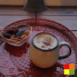 Image of masala chai in a beige mug with brown edges. The mug is on a decorated red tray and next to it is a heart-shaped glass bowl with dried fruits.