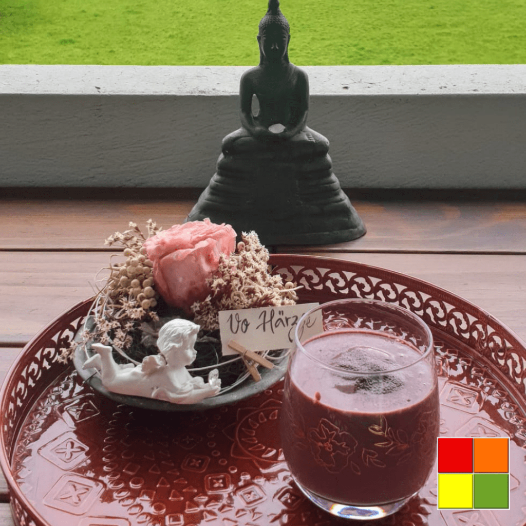 Photo of Cherry Chocolate Smoothie in a glass cup on top of a decorated red tray. Next to the glass there is a flower arrangement, with an angel statue.