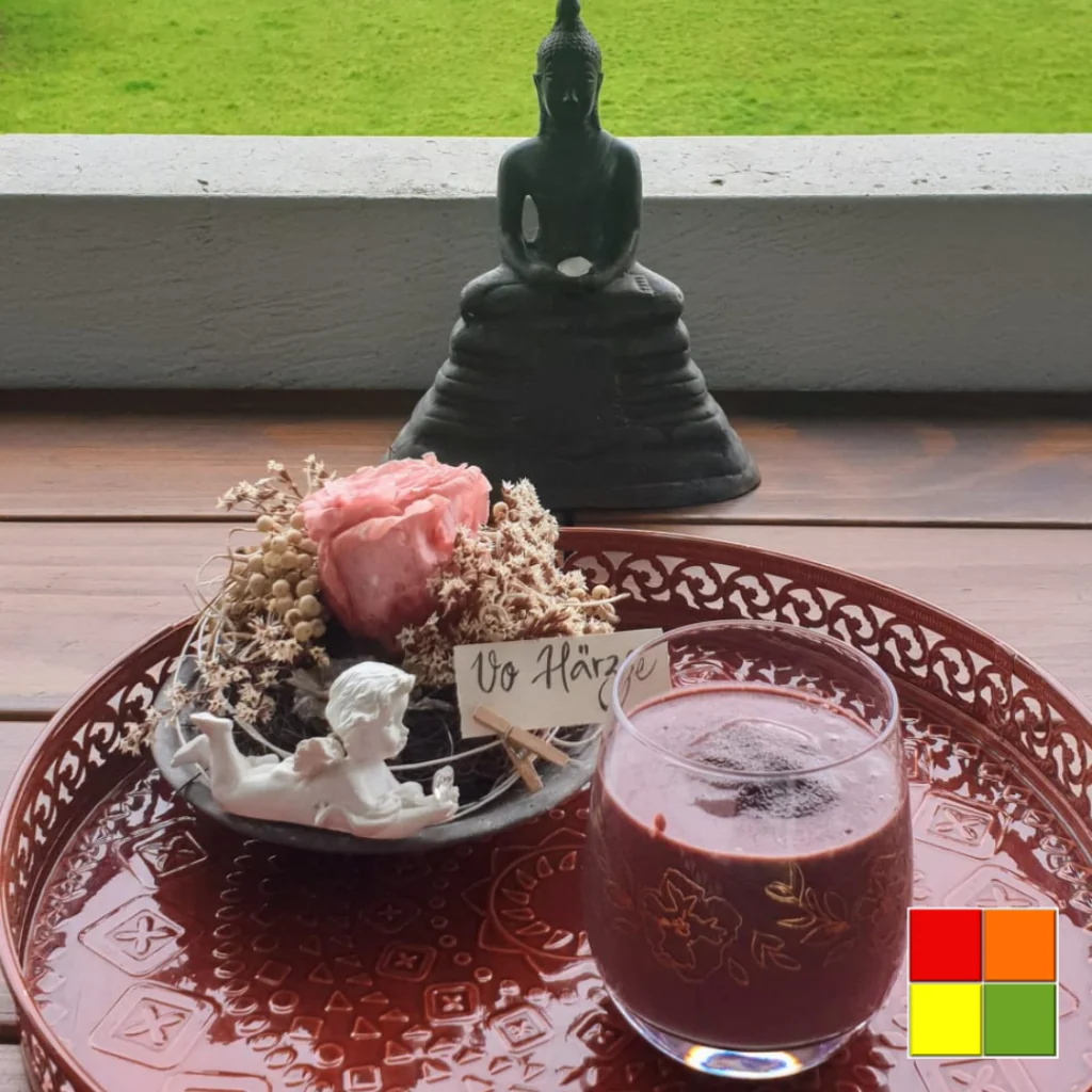 Foto von Kirschschokoladen-Smoothie in einer Glastasse auf einem dekorierten roten Tablett. Neben dem Glas befindet sich ein Blumenarrangement mit einer Engelsstatue.