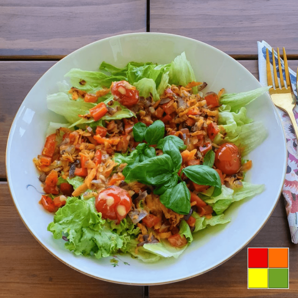 Photo of a white plate of warm salad on a wooden table. Next to the plate is a fork on top of a flowered napkin