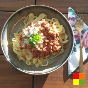 Tagliatelle Vegan Bolognese disajikan dalam mangkuk bergaya pedesaan, dihiasi dengan rempah segar dan keju parmesan vegan, dengan cahaya matahari yang menyoroti hidangan tersebut.