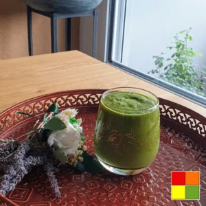 Tulehdusta estävä vihersmoothie tarjoiltuna lasissa koristeellisella tarjottimella kuivattujen kukkien vieressä, ikkunasta tulevan luonnonvalon ympäröimänä.