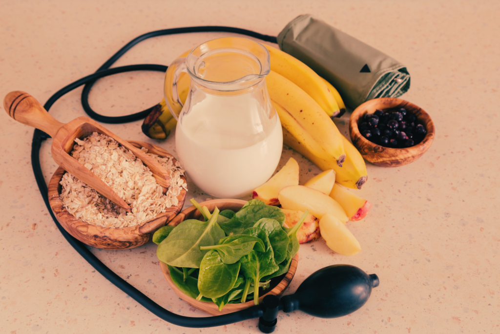 A picture of a sphygmomanometer around healthy foods, including a jug of milk, bananas, a bowl of oats and a bowl of spinach, one with beans and an apple. This picture symbolizes the relationship between Ayurveda and hypertension, in which lifestyle changes are crucial to treatment.