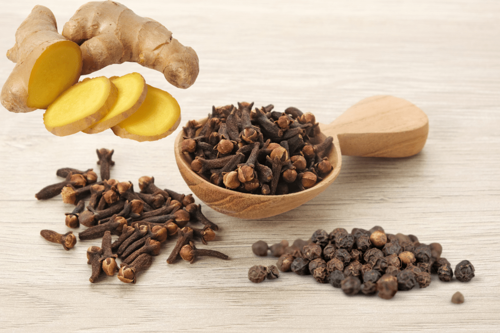 Image of a wooden spoon filled with cloves on a wooden surface, on which there is also sliced ginger and black pepper. Cloves, ginger and black pepper are ingredients in Indian cuisine and in Ayurvedic Remedies for Flu & Throat.