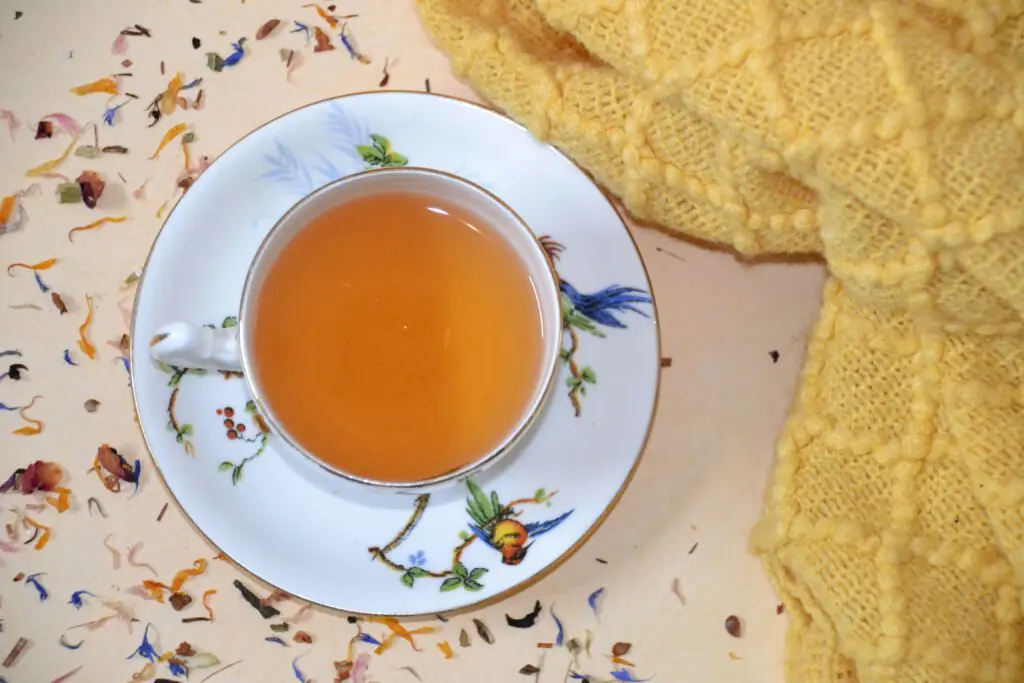 Một tách nước gừng ấm được đựng trong tách trà hoa thanh nhã, đặt trên bàn rắc đầy thảo mộc khô và một chiếc chăn len màu vàng ấm áp — cách làm nước gừng để có sức khỏe tự nhiên.