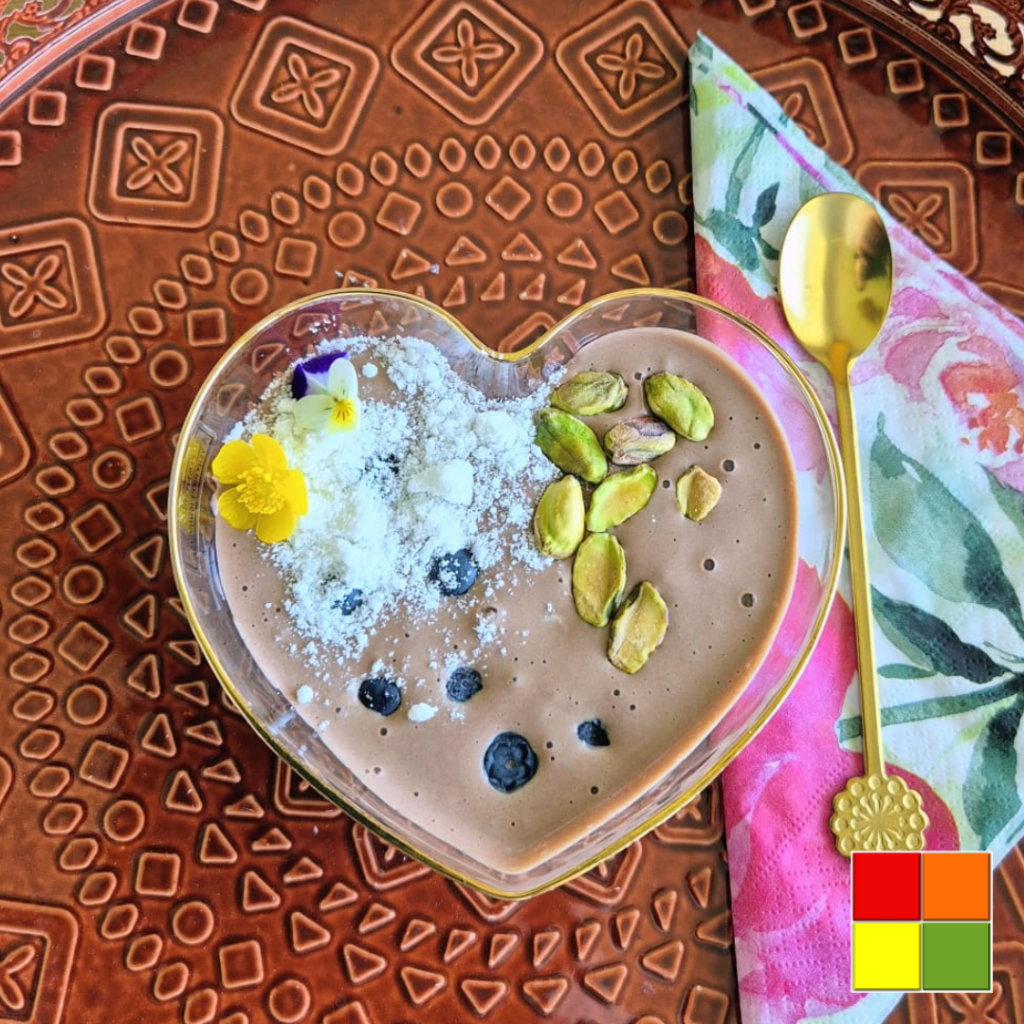 හදවතක හැඩැති අතුරුපස බඳුනක බ්ලූබෙරි, පිස්ටා, පොල් පෙති සහ ආහාරයට ගත හැකි මල් වලින් සරසා ඇති අඩු කාබ් චොකලට් මූස්.