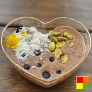 Low Carb Chocolate Mousse garnished with blueberries, pistachios, coconut flakes, and edible flowers in a heart-shaped dessert bowl.